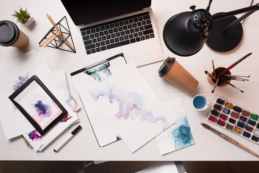 Colorful watercolor paintings on a cluttered desk with art supplies and a laptop.