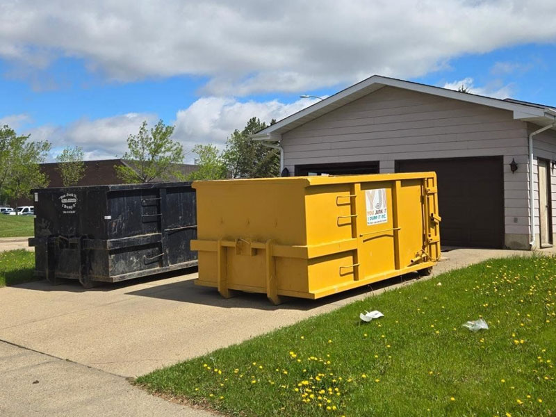 Yellow dumpster on a driveway next to a black junk bin