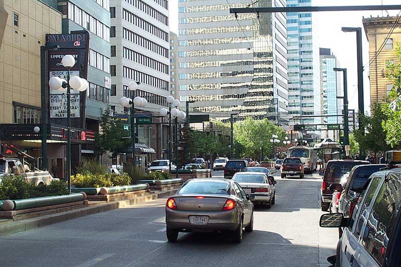 Vehicles parked along a busy city street with tall modern office buildings and trees lining the sidewalk.