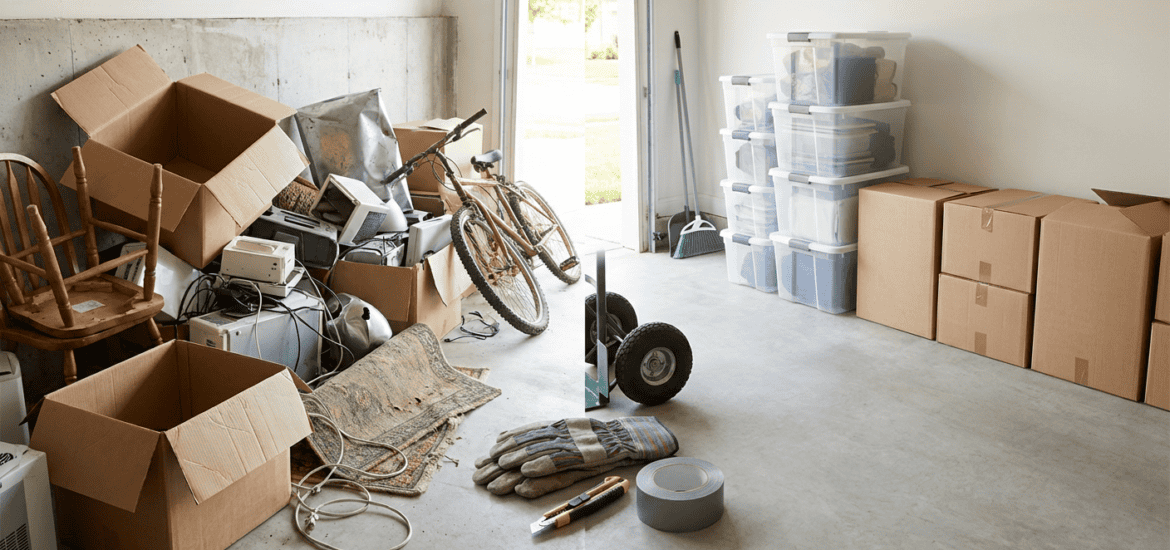 A cluttered storage space filled with open boxes, electronic devices, a bicycle, and various tools, indicating a recent move or storage organization.