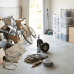 A cluttered storage space filled with open boxes, electronic devices, a bicycle, and various tools, indicating a recent move or storage organization.